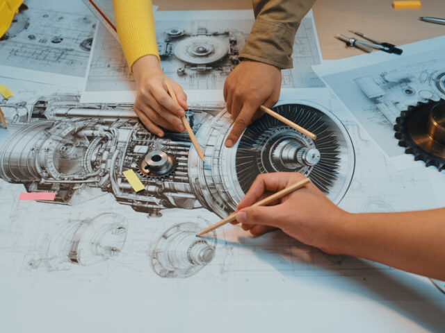 A group of professionals collaborates on a detailed engineering blueprint in a modern workspace, using pencils to highlight critical design elements and showcase teamwork. SACTR